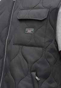 Μαύρο καπιτονέ γιλέκο με μπροστινό φερμουάρ, θήκη στο στήθος και ασημί μεταλλικά στοιχεία. Διαθέτει πατς με το λογότυπο Guess Jeans στην αριστερή πλευρά.