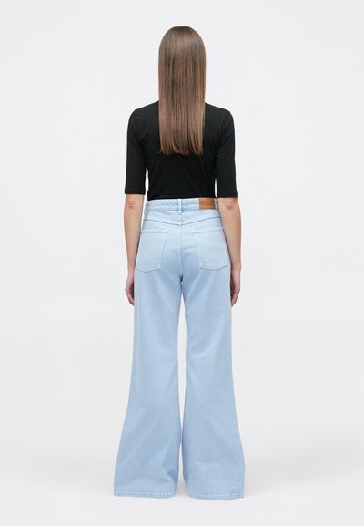 Γυναίκα με ίσια μακριά μαλλιά, ντυμένη με μαύρο τοπ και ανοιχτόχρωμο γαλάζιο ψηλόμεσο τζιν παντελόνι με φαρδιά μπατζάκια, που στέκεται γυρισμένη με την πλάτη.