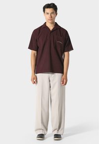 Camicia cortas maniche color marrone testo con colletto, dotata di tasca frontale e dettagli ricamati, abbinata a pantaloni larghi beige chiaro.