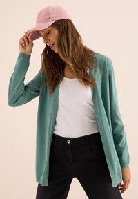 Žena s růžovou čepicí, tyrkysovým kardiganem, bílým topem a černými džíny, naklání hlavu dolů, zatímco drží okraj čepice.