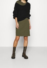 Czarny sweter z dzianiny z podwiniętymi rękawami, nałożony na oliwkową sukienkę. Modelka nosi ciemnozielone botki za kostkę z grubymi podeszwami.