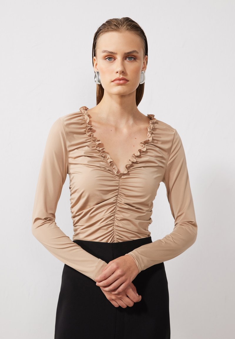 Beige langermet topp med volangkant, samlet stoff i midten og glatt tekstur. Flott sammen med et svart skjørt, modellen poserer direkte.