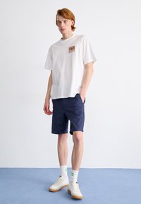 T-shirt bianca in cotone con un logo grafico quadrato arancione e marrone, abbinata a pantaloni navy e sneakers bianche con suola in gomma chiara.