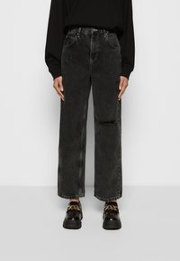 Jeans de ganga pretos de perna larga com cintura alta, apresentando um detalhe desgastado numa perna e costura destacada. Combinados com sapatos pretos robustos.