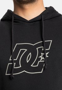 Sudadera negra de algodón, con un gran logo gris de DC en la parte delantera. Incluye cordones en la capucha y dobladillo acanalado.