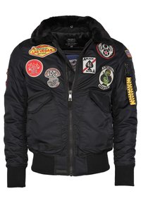 Svart bomberjacka med fuskpälskrage, dragkedja och flera lappar inklusive logotyper och mönster. Har ribbade ärmslut och nederkant.