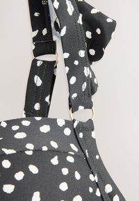 Svart BH-band med vita prickar, utrustat med en justeringsring i guldfärgad metall och texturerat tyg. Närbild på bandets detaljer.