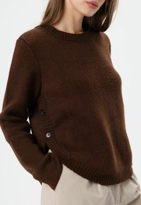 Brauner Strickpullover mit Rundhalsausschnitt, strukturiertem Stoff und seitlichen Knöpfen. Verfügt über gerippte Säume und einen lockeren Schnitt. Kombiniert mit hellen Hosen.