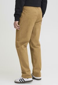 Tan šīfera kokvilnas bikses ar taisnu kāju dizainu, kas izceļas ar aizmugurējo kabatu, redzamu šuvi un valkātas kopā ar baltām sporta kurpēm un melnām svītrām.