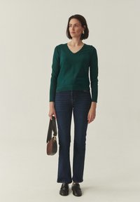 Pull vert à col en V, poignets côtelés, coupe ajustée ; jean évasé bleu foncé ; chaussures noires ; portant un petit sac bandoulière marron.