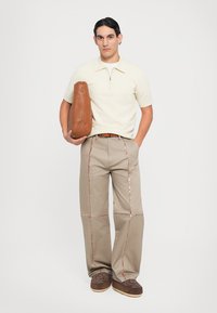 Korte crèmekleurige polo met rits, beige broek met pailletdetails, bruin tas en donkerbruine schoenen met een bloemenpatroon.