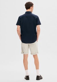 Marinblå kortärmad skjorta med en avslappnad passform, tillsammans med ljusbeige uppvikta shorts och svarta skor, sedd bakifrån.