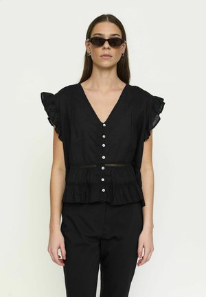 Nő fekete fodros, rövid ujjú blúzban fehér gombokkal, fekete nadrágban, karika fülbevalóval és sötét napszemüvegben, egyszerű háttér előtt.