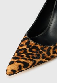 Leparda raksta augstpapēžu kurpe ar spicajiem pirkstiem, apdarināta ar melnas ādas akcentiem un strukturētu, kažokādas līdzīgu virsmu brūnos un melnos toņos.