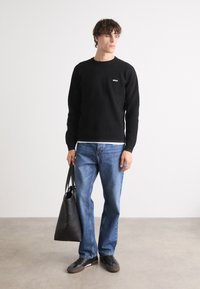 Czarny bawełniany sweter z okrągłym dekoltem i małym logotypem, w połączeniu z niebieskimi dżinsami. Modelka trzyma czarną skórzaną torbę i ma na sobie szare sneakersy.