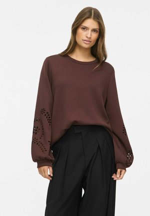 Femme portant un haut marron à manches longues avec des découpes à motifs sur les manches et un pantalon plissé noir taille haute sur un fond uni.