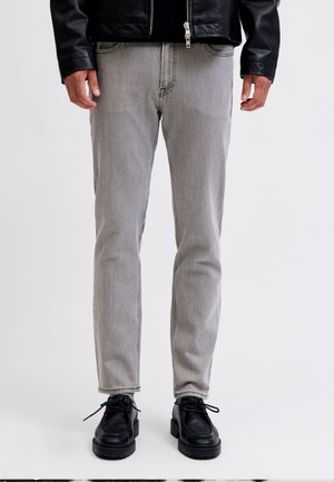 Personne portant un jean gris clair, une veste en cuir noire et des chaussures noires à lacets, debout devant un fond uni.