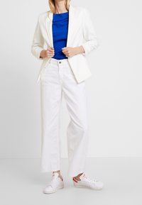 Vit skräddarsydd blazer, blå topp, vita vida byxor och vita sneakers med röda accenter. Mjukt tyg, rena linjer och modern design.