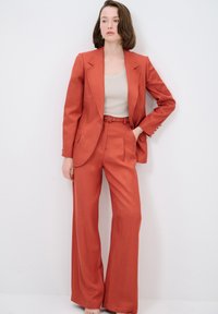 Rust-colored suit featuring a tailored blazer with pockets and detailed stitching, paired with high-waisted wide-leg trousers. Smooth fabric texture.