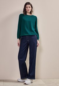 Teal texturerad långärmad top i kombination med mörka vida jeans och vita sneakers. Toppen har en enkel halsringning och rynkade ärmar.