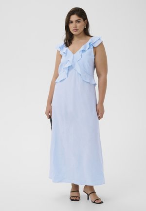 Žena u svjetloplavoj maksi haljini bez rukava s naboranim V-izrezom i crnim sandalama s remenčićima, stoji ispred jednobojne sive pozadine.