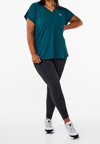 Teal v-hals t-shirt i let materiale, parret med sorte leggings. Har korte ærmer og et subtilt logo på brystet.