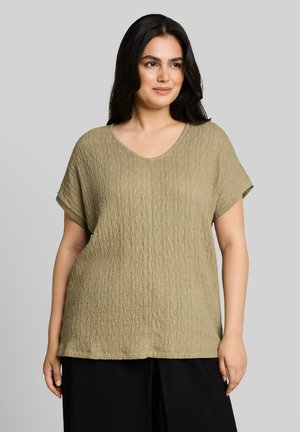 Femme aux longs cheveux foncés portant un haut ample texturé vert olive à manches courtes et un pantalon noir, debout devant un fond gris uni.