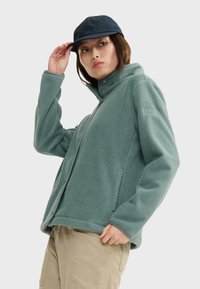 Fleece jacket in muted teal with a high collar, zip closure, and snap buttons. Features textured fabric and an embroidered logo on the sleeve.