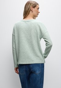 Jasnozielony sweter teksturowany o luźnym kroju, z szerokim dekoltem i opadającymi ramionami, zestawiony z niebieskimi dżinsami, widok z tyłu.