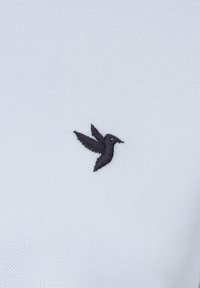 Polo in tessuto azzurro chiaro con un piccolo logo ricamato di un colibrì nero sul lato sinistro. Superficie testurizzata.
