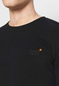Schwarzes Baumwoll-T-Shirt mit rundem Ausschnitt und kurzen Ärmeln. Verfügt über ein kleines, besticktes "SuperDry"-Logo in dunklem Faden und einem orangefarbenen Akzent.