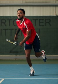 Camisa atlética roja con acentos blancos, pantalones cortos azul marino y zapatillas de tenis azules. Jugador sosteniendo una raqueta en una cancha cubierta.