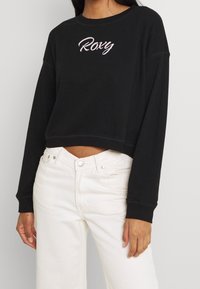 Felpa nera corta con ricamo in rosa della scritta "Roxy". Presenta maniche lunghe e una vestibilità morbida. Abbinata a jeans bianchi a vita alta.