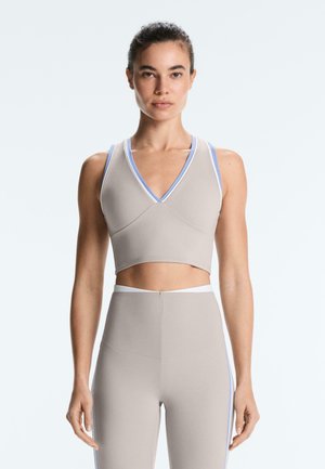 Ženska v bež brezrokavniku in visokim pasom nogavicah z modrim in belim robom, stoji na svetlem ozadju.