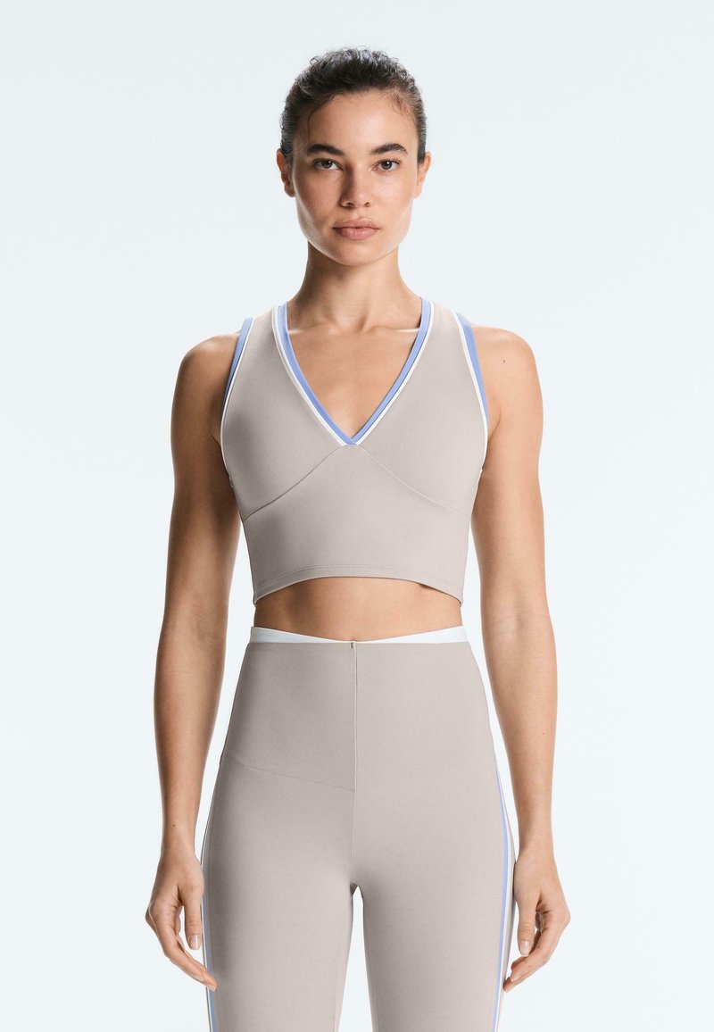 Femme portant un crop top beige sans manches et des leggings taille haute avec une bordure bleue et blanche, debout sur un fond clair.
