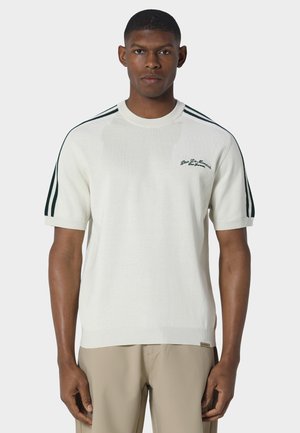 Mand, der står fremadrettet iført en hvid kortærmet sweater med mørke striber på skuldrene og beige bukser.