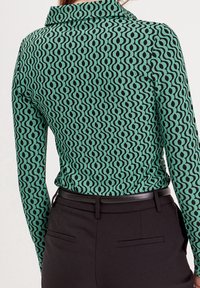 Femme portant un haut vert ajusté à manches longues avec un motif géométrique noir, rentré dans un pantalon noir taille haute avec une ceinture noire fine.