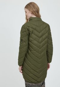 Olivengrøn pufferjakke med diagonal chevron-syning, høj krave og sort logo-patch på ærmet. Letvægts, quiltet tekstur, knælang.