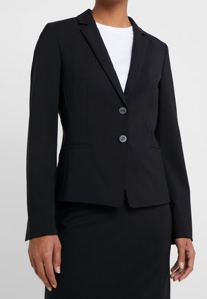 Kobieta nosząca dopasowaną czarną marynarkę na dwa guziki na białą bluzkę i dopasowaną czarną spódnicę, stojąca na jednolitym tle.