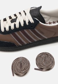 Ruskeat Adidas Samba -urheilukengät mustilla mokka-yksityiskohdilla, valkoisella sahalaitapistokkeella, kermaiset nauhat sekä kaksi kierrettyä ruskeaa ja valkoista varanauhaa.