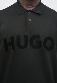 Sötétzöld pamut póló texturált felülettel. Háromgombos gombsorral és a mellrészen feltűnő fekete "HUGO" logóval.