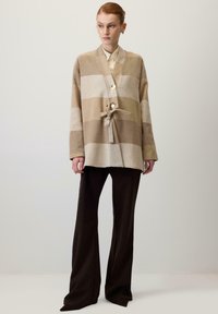 Bēšs un krēmkrāsas svītrains kardigans ar aizdari, kas saistīta ar lentu, ar šalles apkakli un lielām pogām, kopā ar tumši brūnām platām biksēm.