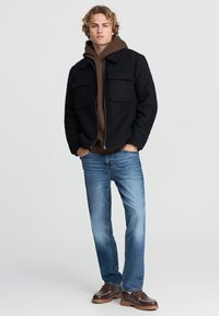 Giacca in pile nera con due tasche anteriori sopra un hoodie marrone, abbinata a jeans blu e scarpe in pelle marrone con dettagli di cucitura.