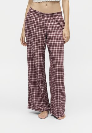 Femme portant un pantalon de pyjama large à carreaux bordeaux et blancs, debout pieds nus sur un fond uni.