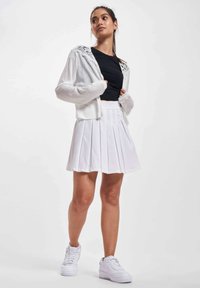 Weißer, kurz geschnittener Pullover mit Spitzendetails, kombiniert mit einem schwarzen, figurbetonten Oberteil und einem weißen, plissierten Rock. Abgerundet mit weißen Sneakers.