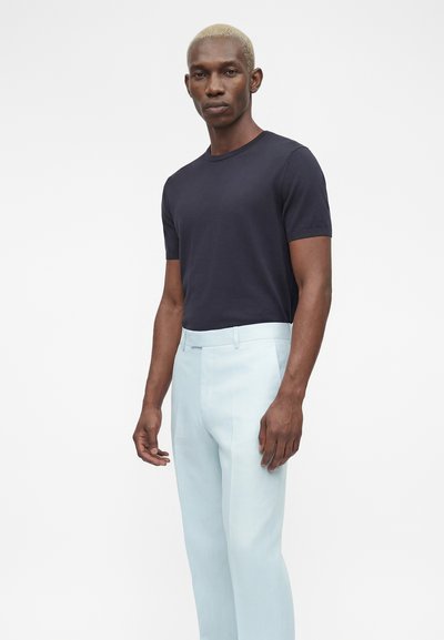 Άνδρας με κοντά ξανθά μαλλιά, που φοράει ένα εφαρμοστό μαύρο κοντομάνικο πουκάμισο και ανοιχτό μπλε ραμμένα παντελόνια, πάνω σε απλό λευκό φόντο.