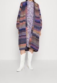 Sweter robiony na drutach w wielokolorowe paski (fioletowy, pomarańczowy, niebieski) z długimi rękawami, otwarty z przodu, noszony na fioletowej sukience w kwiaty i białych botkach.
