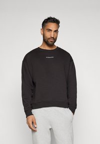 Μαύρη μπλούζα με στρογγυλή λαιμόκοψη και ριμπ μανσέτες, με μικρό λευκό κείμενο, συνδυασμένη με γκρι φόρμες. Απλός, �άνετος σχεδιασμός.