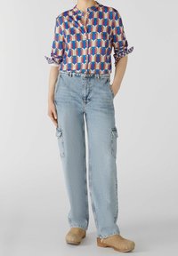 Blusa a fantasia multicolore con colletto, abbinata a jeans cargo azzurri chiari con tasche laterali e zoccoli beige.