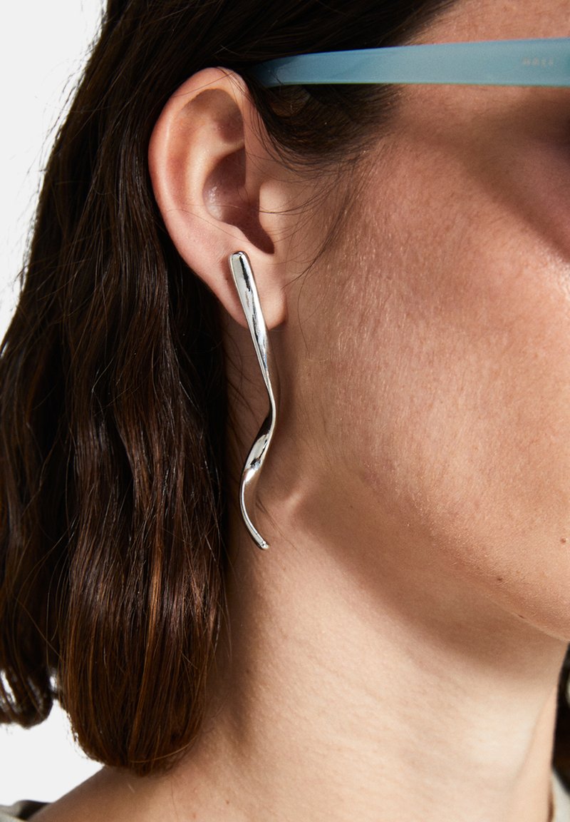 Silverfärgade metallörhängen med en böjd, vågig design. Slät yta med reflekterande yta. Bärs på vänster öra.